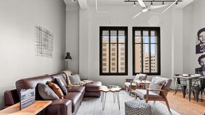 Una sala de estar moderna con un sofá de cuero, una mesa de centro de madera y un reposapiés. Hay dos ventanas con marcos negros, vistas a la ciudad y una gran obra de arte abstracta en la pared.