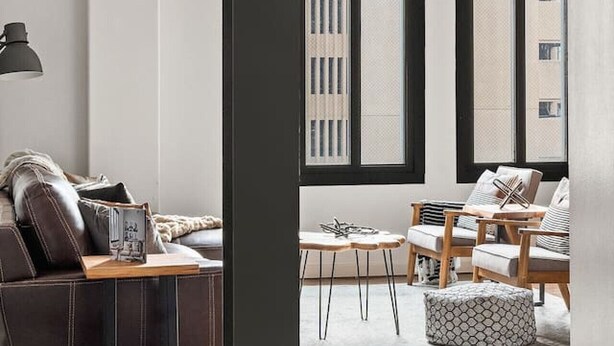 Una sala moderna con un sofá de cuero, una mesa de centro de madera, y un comedor con sillas y una mesa. Amplios ventanales ofrecen luz natural y vistas de la ciudad.