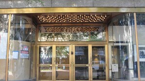 Entrada de edificio con puertas de cristal, un letrero de advertencia y un aplique decorativo sobre las puertas.