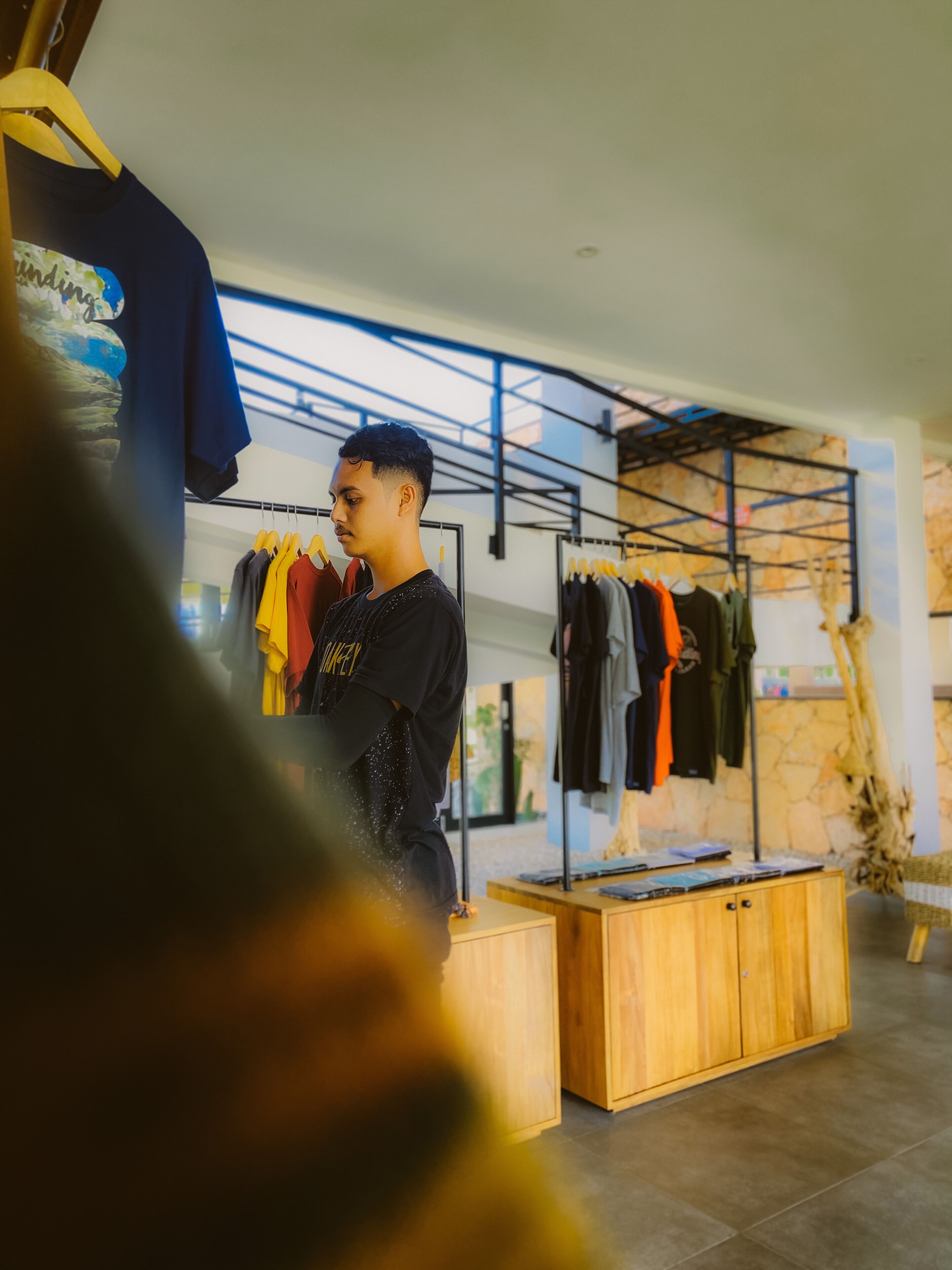 Une personne se tient dans un espace de vente moderne, avec des présentoirs à vêtements et une table d’exposition en bois.
