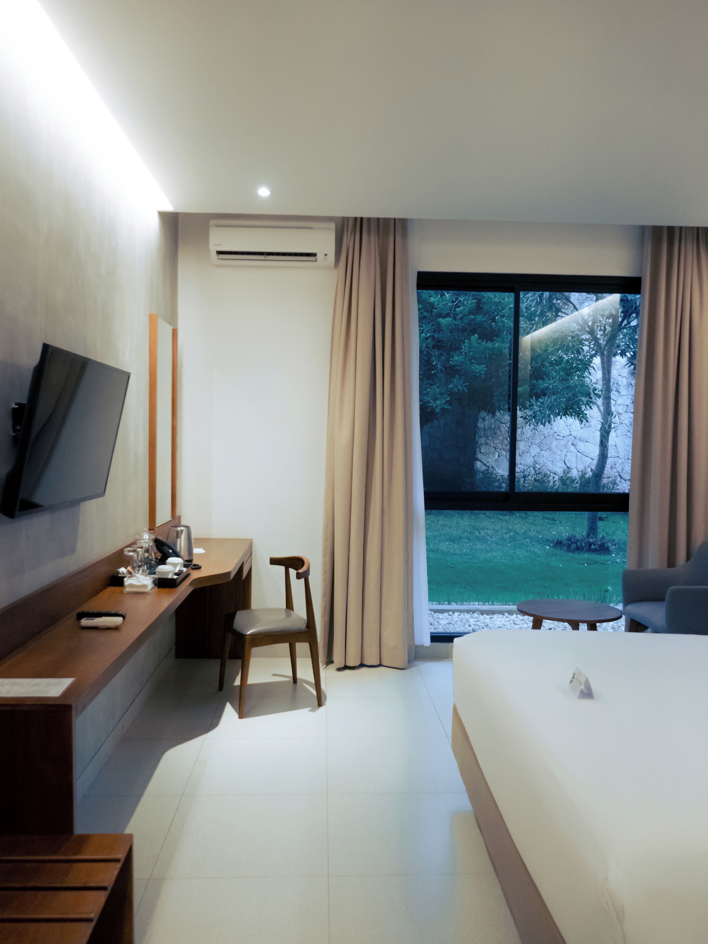 A modern hotel room with a bed, a desk, a chair, a TV, and a large window with a view of greenery.