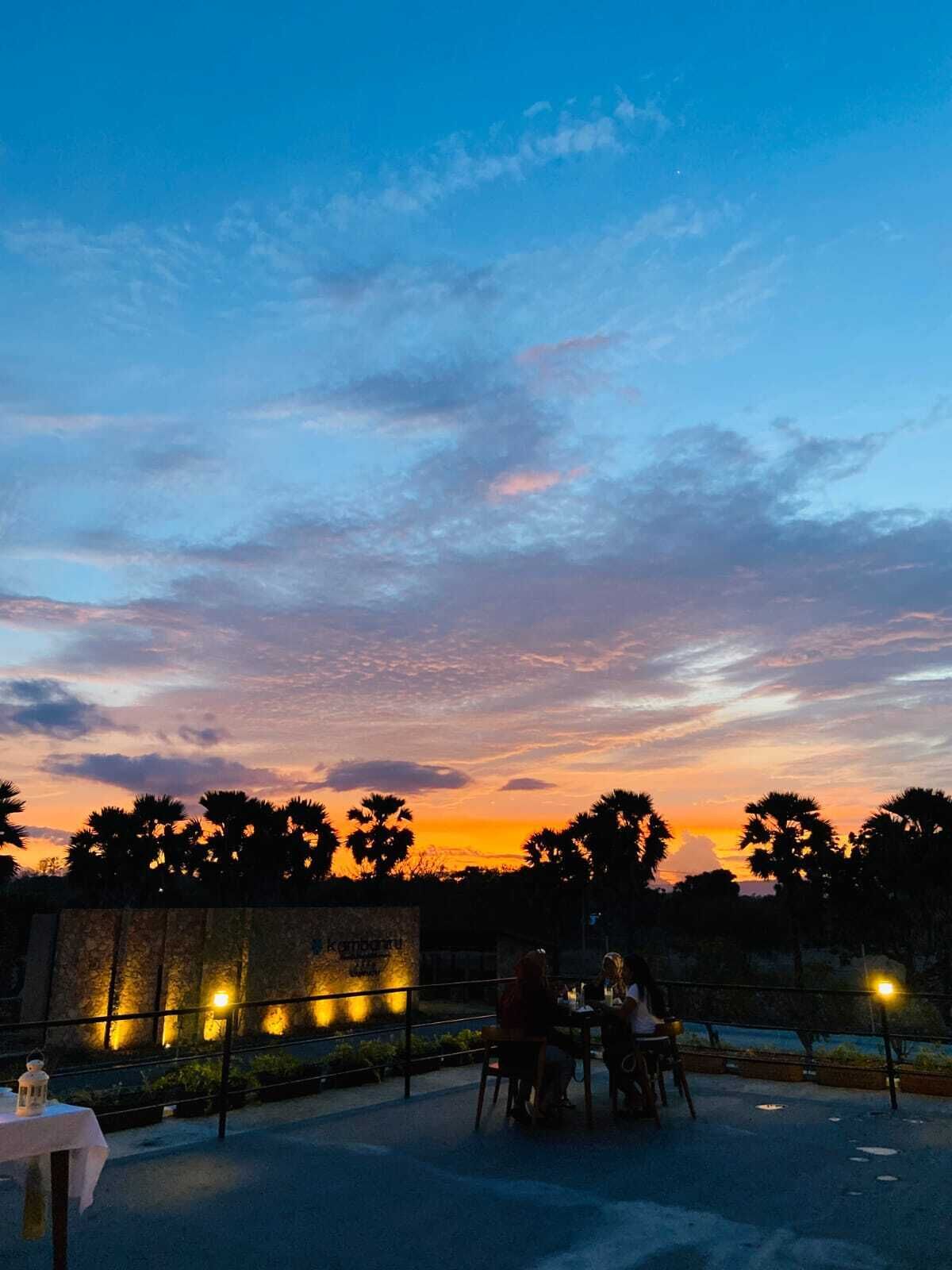 An outdoor dining area with a sunset view, a stone wall with lights, and a group of people dining.