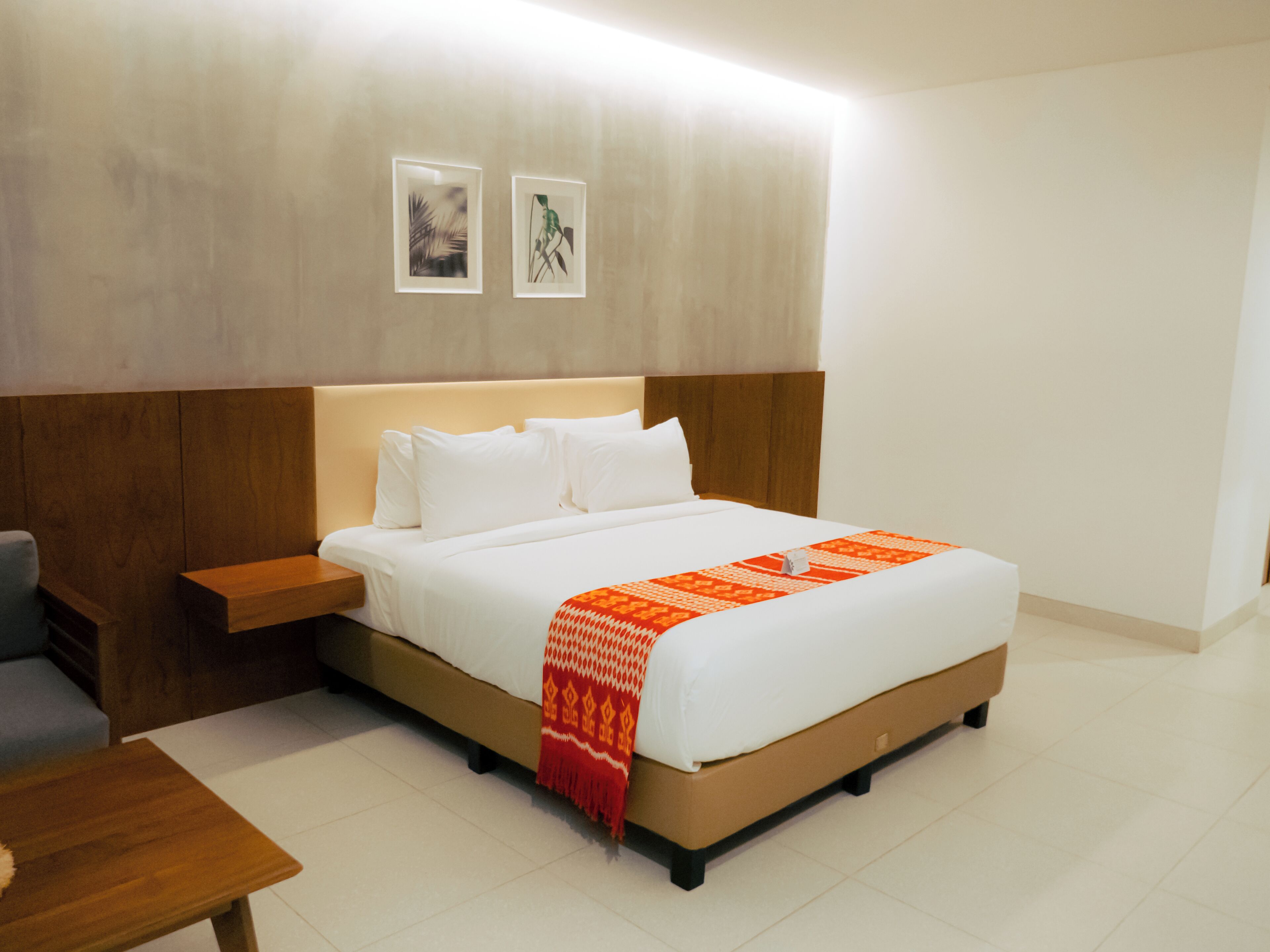 A hotel room with a large bed, a wooden nightstand, a small table, and two framed pictures on the wall.