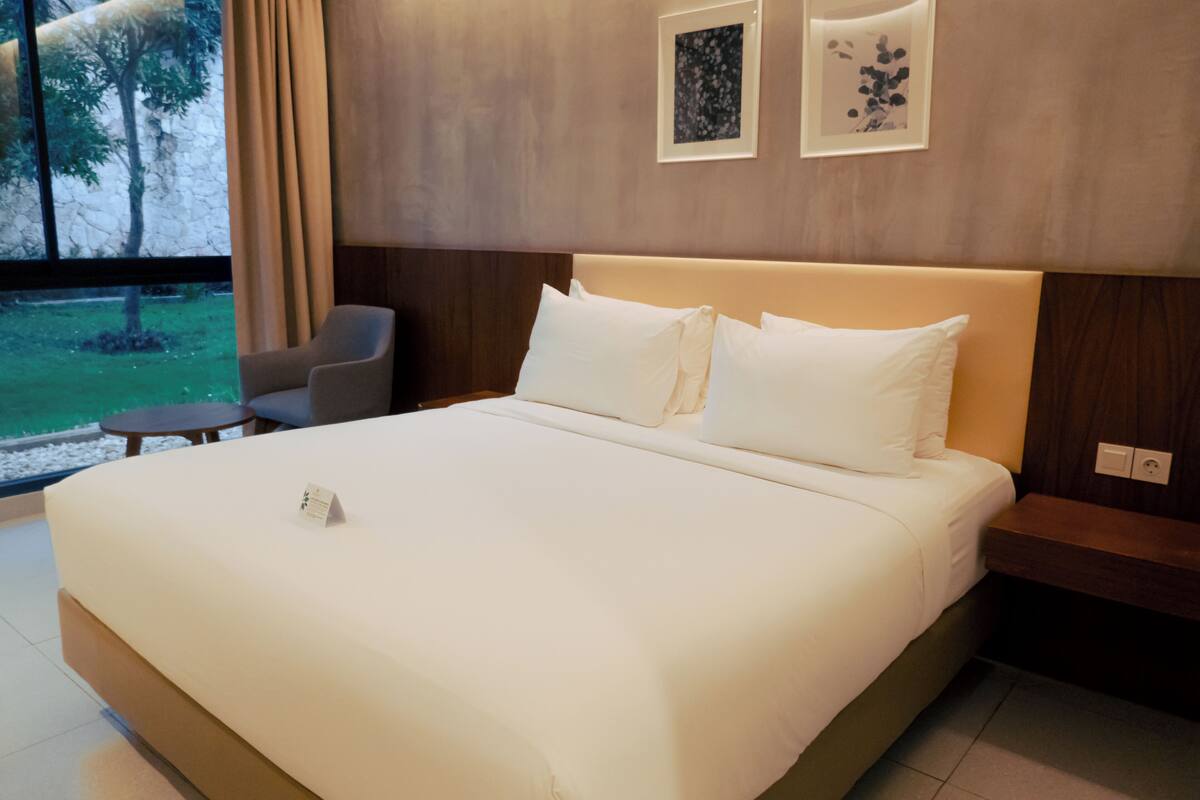 Ein Hotelzimmer mit einem großen Bett, einem kleinen Tisch, einem Sessel und Blick ins Freie.