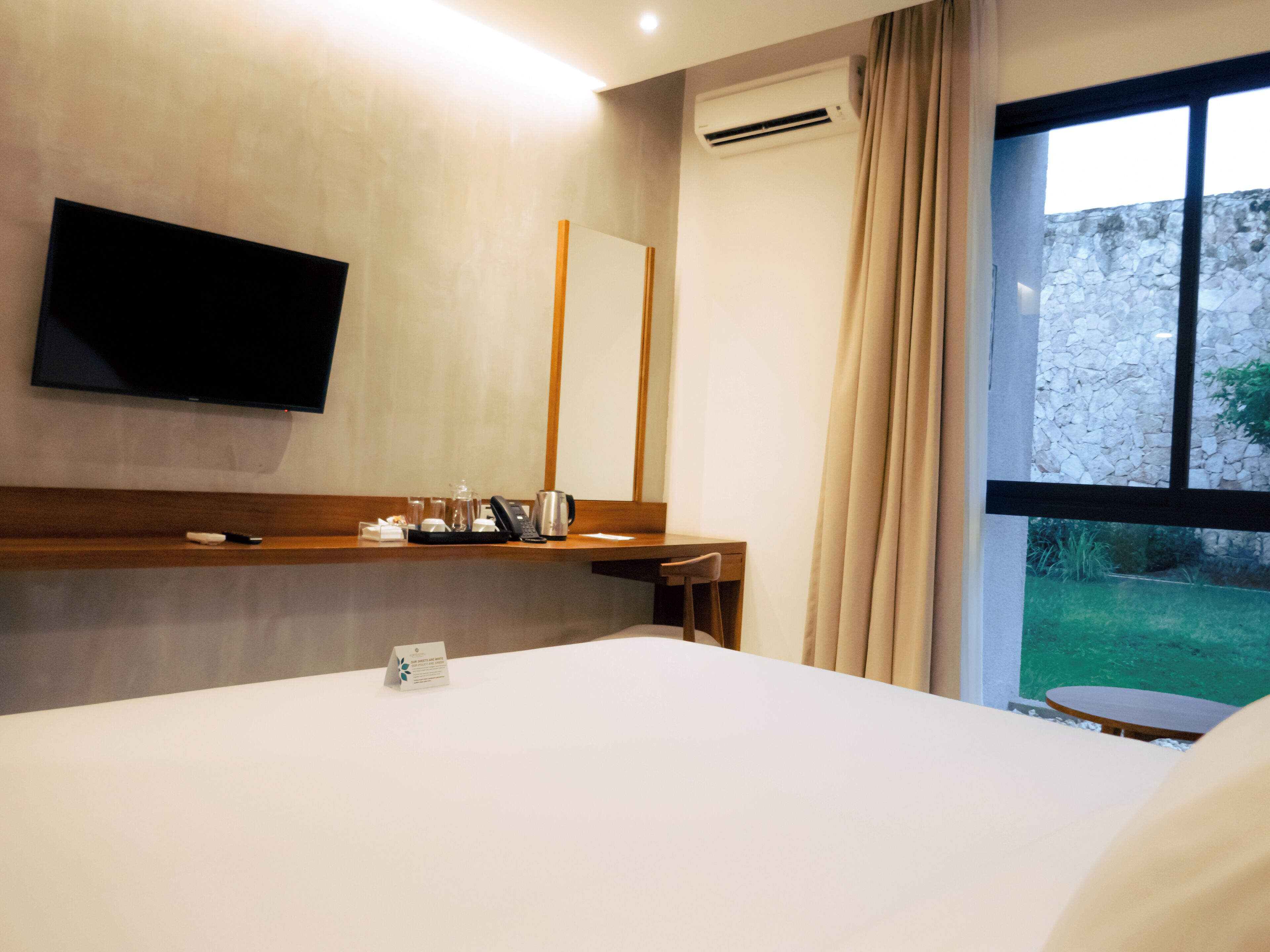 Una habitación de hotel con cama, televisor montado en la pared, escritorio con teléfono y ventana con cortinas.