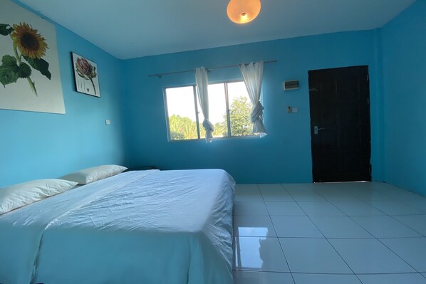 Una habitación con cama, ventana con cortinas, puerta y un cuadro de un girasol en la pared.