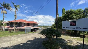 A large two-story house with a red roof, surrounded by a fence and palm trees. A signboard reads "RB Homestay & Farms". - Hotel O Rb Homestay & Farms (Kuching)