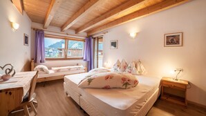 Une chambre avec un plafond en bois, un lit avec une literie à motifs floraux, une table de chevet, un bureau avec une chaise et une fenêtre donnant sur une montagne.