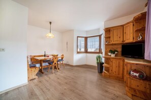 A room with a wooden dining table, chairs, a hanging light fixture, a television, and wooden cabinets.