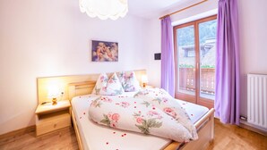 Ein Schlafzimmer mit einem Bett, einem Nachttisch, einer Wandleuchte, einem Fenster mit violetten Vorhängen und Blick auf ein Gebäude.