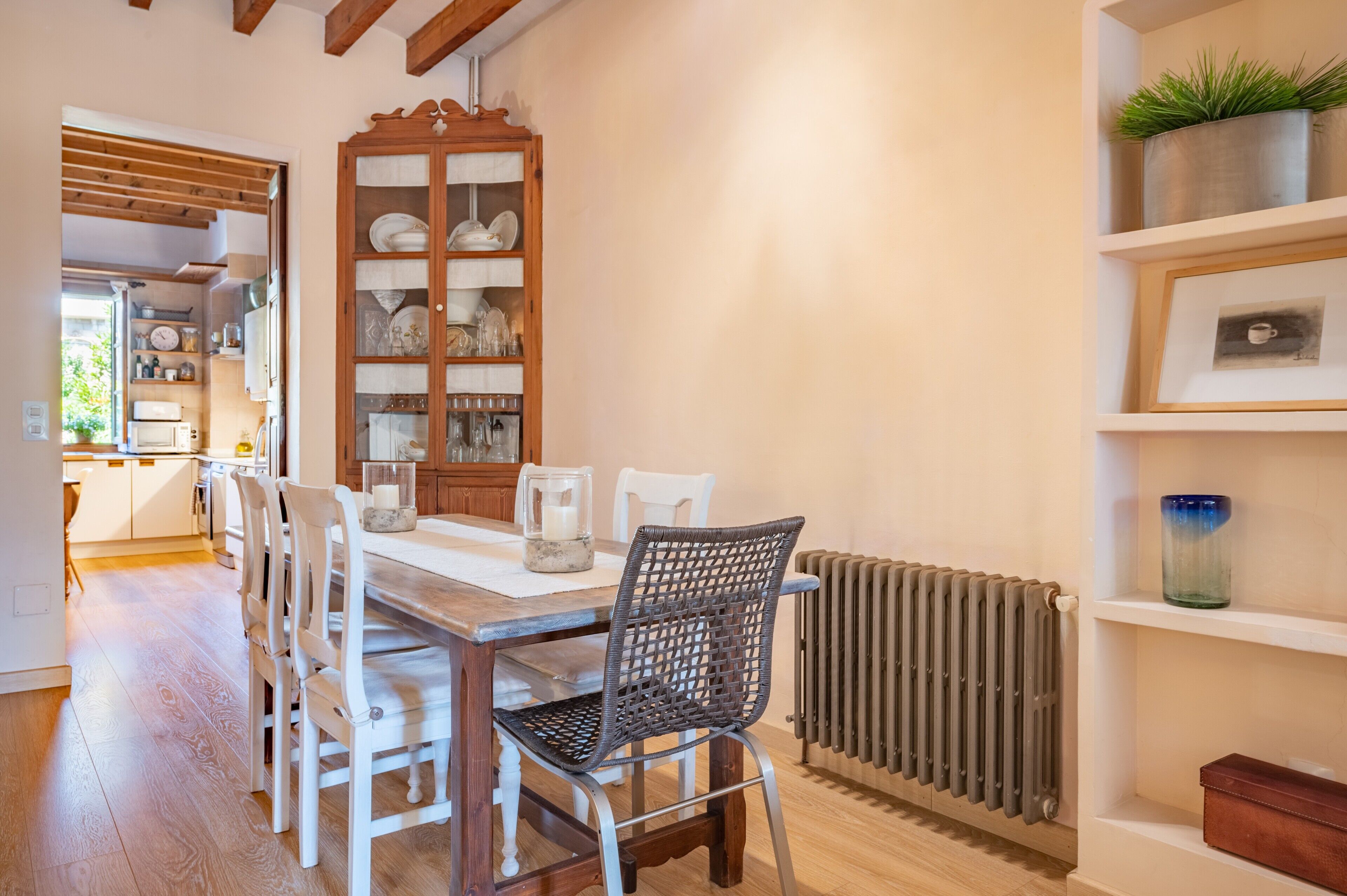 Un comedor con mesa de madera, sillas blancas, una silla de mimbre, un armario con vajilla, un radiador y estanterías con plantas en macetas.