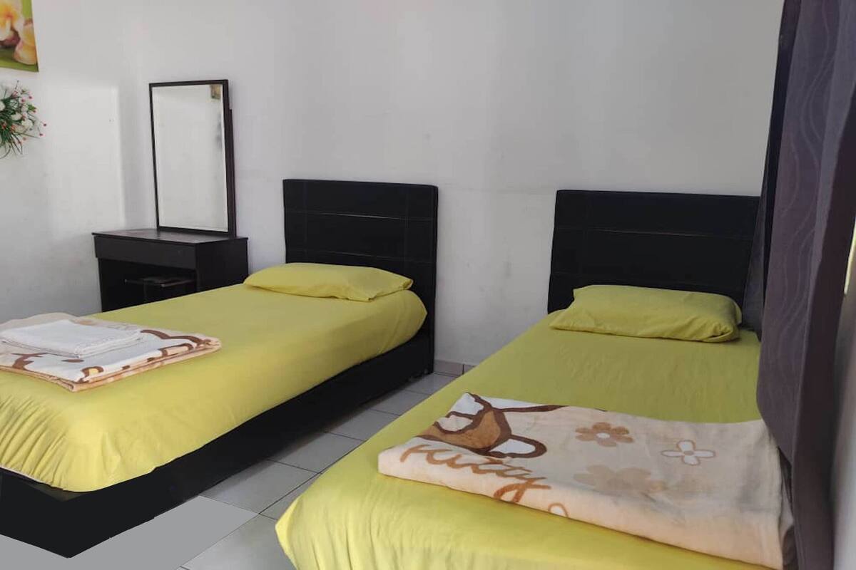Ein Hotelzimmer mit zwei Betten, jeweils mit einer gelben Tagesdecke und einem Kissen. Es gibt einen Nachttisch mit einem Spiegel und einer kleinen Pflanze.