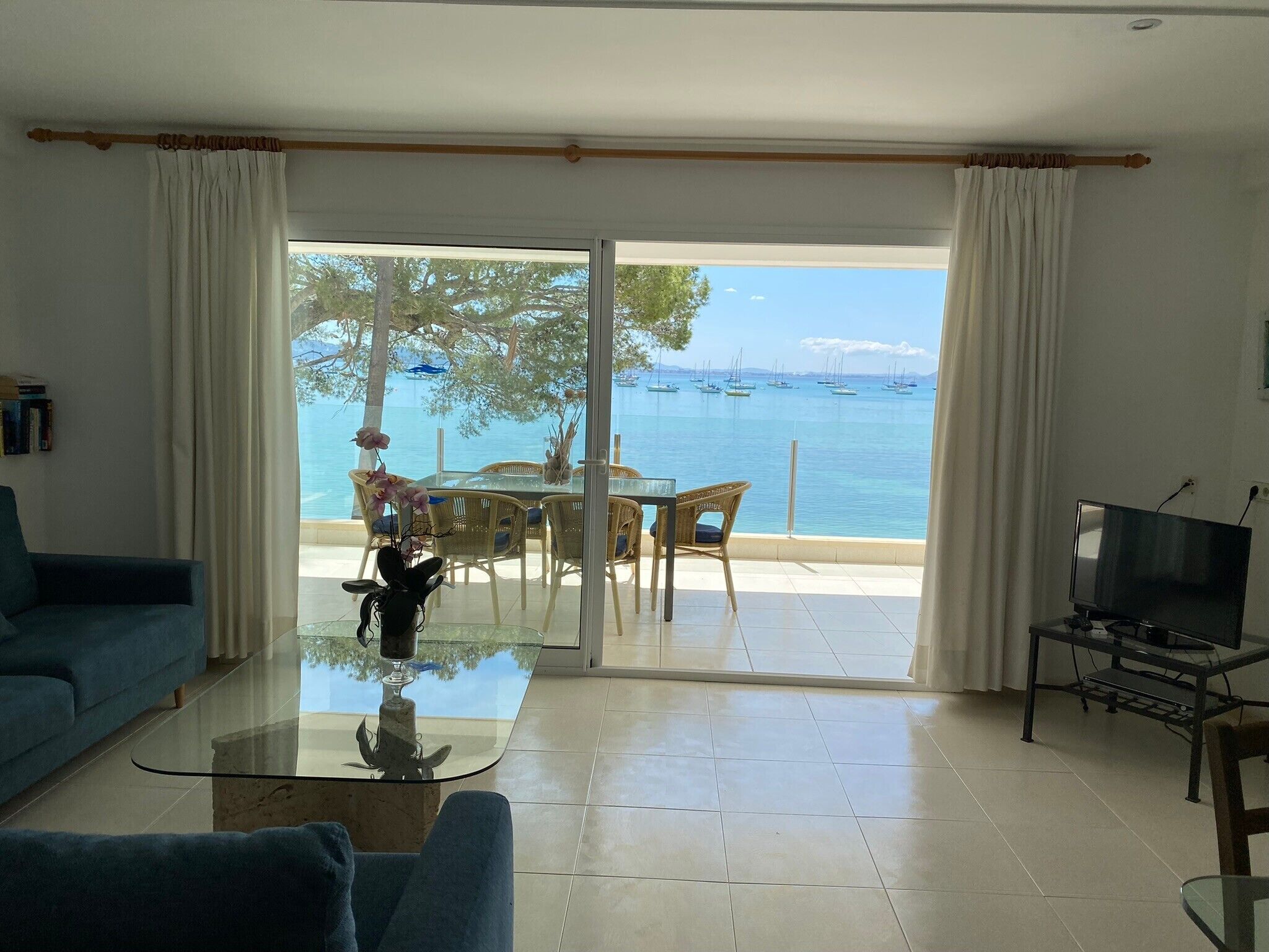Ett vardagsrum med ett soffbord i glas, en soffa, en tv och utsikt över havet med båtar.