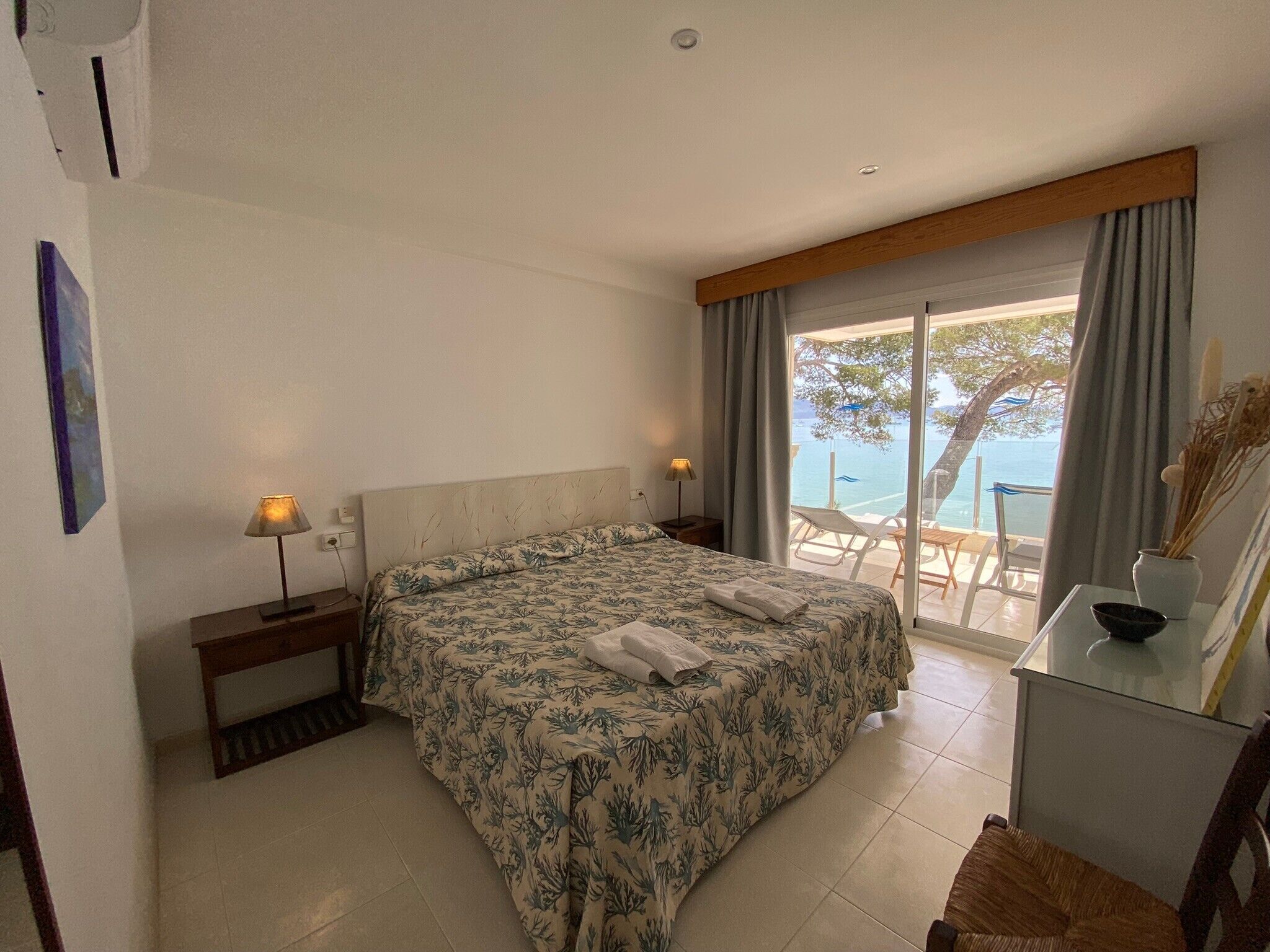 A bedroom with a bed, bedside lamps, a desk, and a view of the sea.