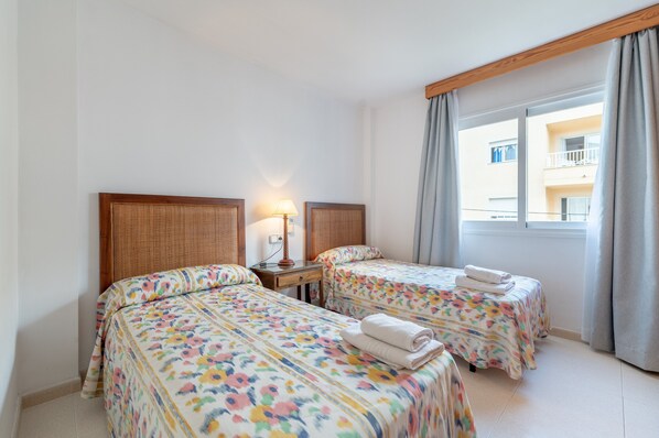 A hotel room with two beds, a nightstand, a lamp, and a window with curtains. - Holiday Home Catalina 2A Tres Dormitorios with Sea View, Wi-Fi & Terrace (Puerto Pollensa)