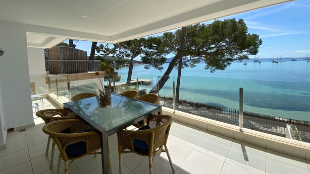 Een balkon met een glazen tafel en rieten stoelen, met uitzicht op een heldere zee en boten.
