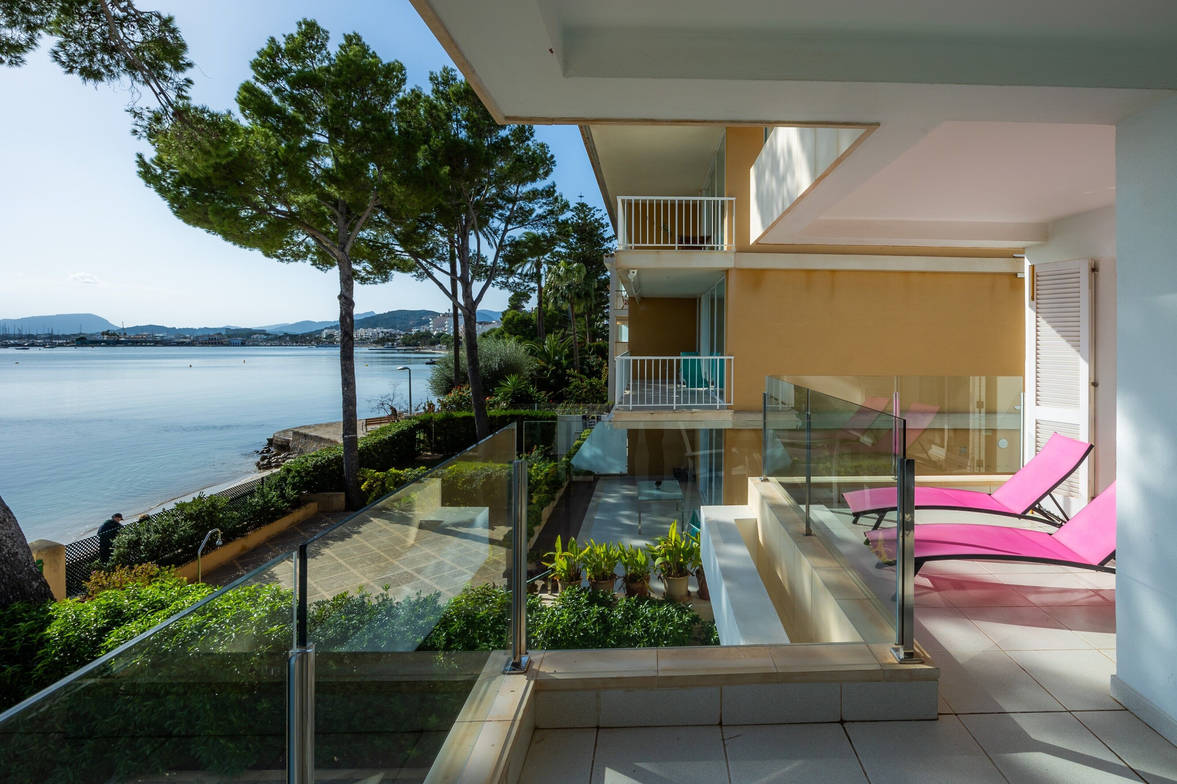 Un apartamento moderno con balcón de vidrio con vistas al agua, un balcón en el segundo piso y una terraza con plantas en macetas.