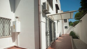 Un estrecho pasillo exterior con suelo de baldosas, pared blanca, portón metálico y toldo blanco.
