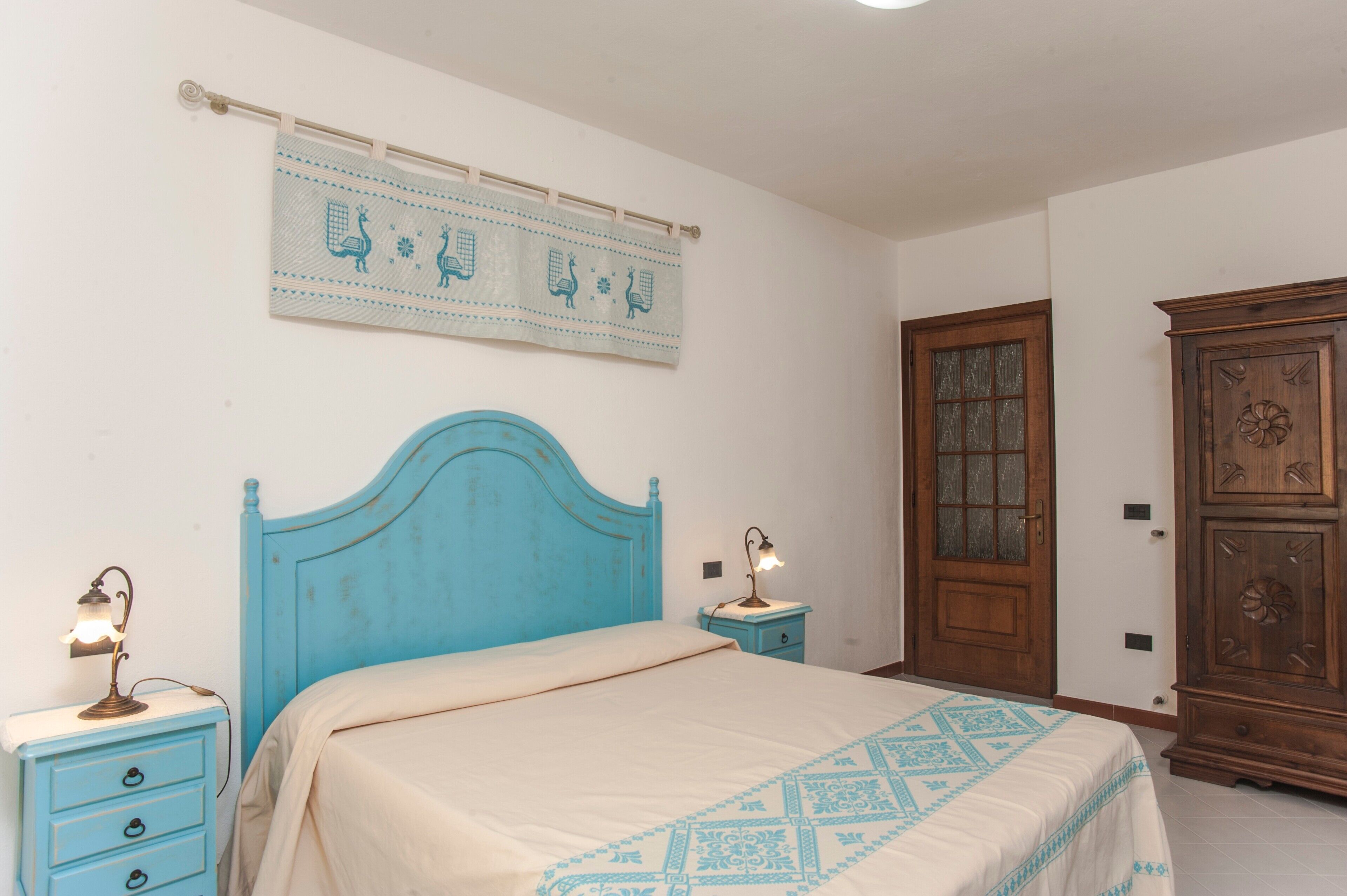 Un dormitori amb un panell blau al capçal, un llit amb un edredó estampat, una meseta de nit amb una làmpada, un armari de fusta i un penjoll a la paret.