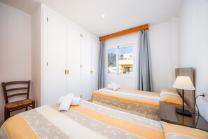 Une chambre d’hôtel avec deux lits, une chaise en bois, une table de chevet avec une lampe et une fenêtre avec des rideaux.