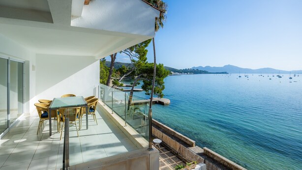 Un balcon doté d’une balustrade en verre donnant sur une étendue d’eau, une table avec des chaises et une vue sur des montagnes lointaines.