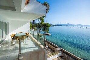 Un balcon doté d’une balustrade en verre donnant sur une étendue d’eau, une table avec des chaises et une vue sur des montagnes lointaines.