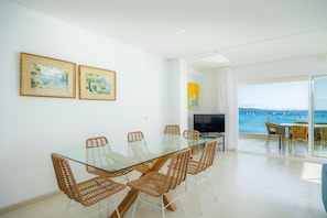Zona pranzo moderna con tavolo in vetro e sedie in vimini, una televisione e vista sul mare.