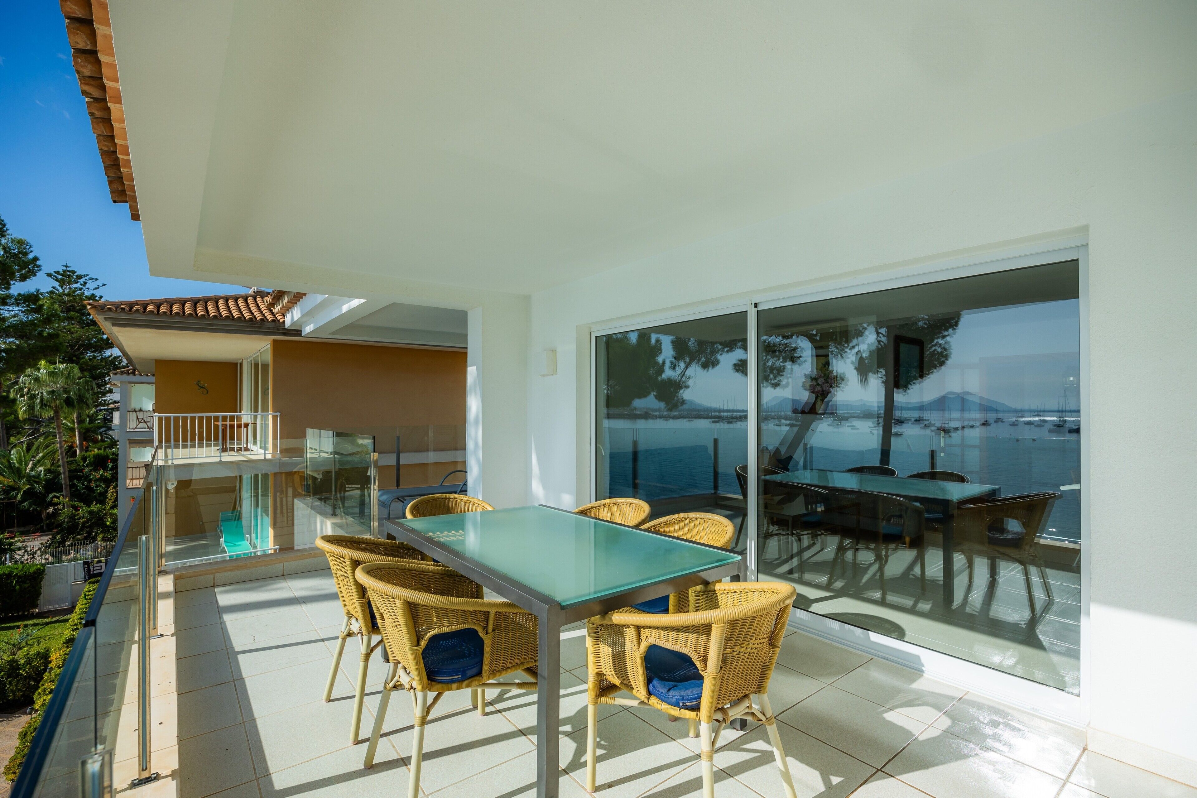 A sunlit patio with a glass table and wicker chairs, overlooking a scenic view.
