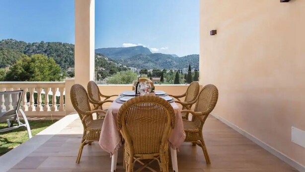 Un balcon agrémenté de fauteuils en osier et d’une table dressée pour le repas, offrant une vue sur un paysage montagneux.