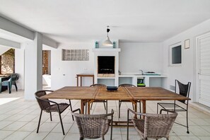 A dining area with a wooden table and wicker chairs, a white wall with a window, and a stone wall partially visible.