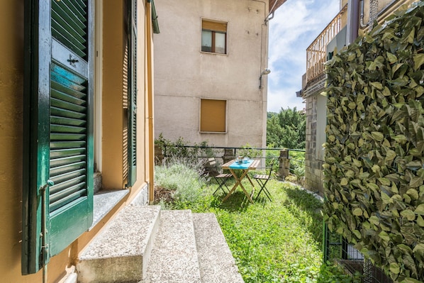 A courtyard with steps leading to a wooden table and chairs, surrounded by greenery and a building with a balcony.