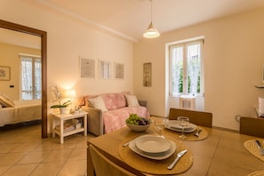 A cozy living room with a sofa, a dining table set for two, and a small side table with a lamp and a plant.