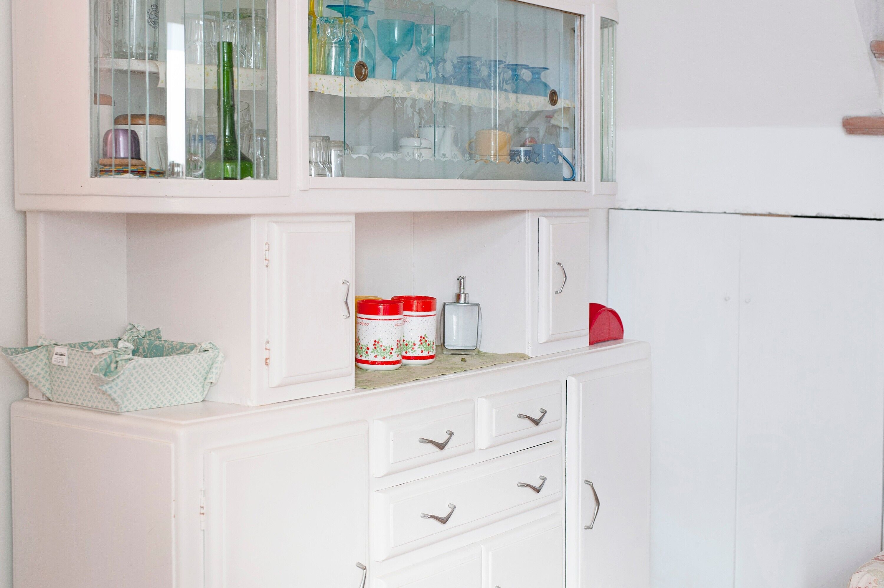 Une cuisine avec des meubles blancs, une étagère supérieure à façade en verre où sont rangés de la vaisselle, et un objet rouge sur l’étagère inférieure.