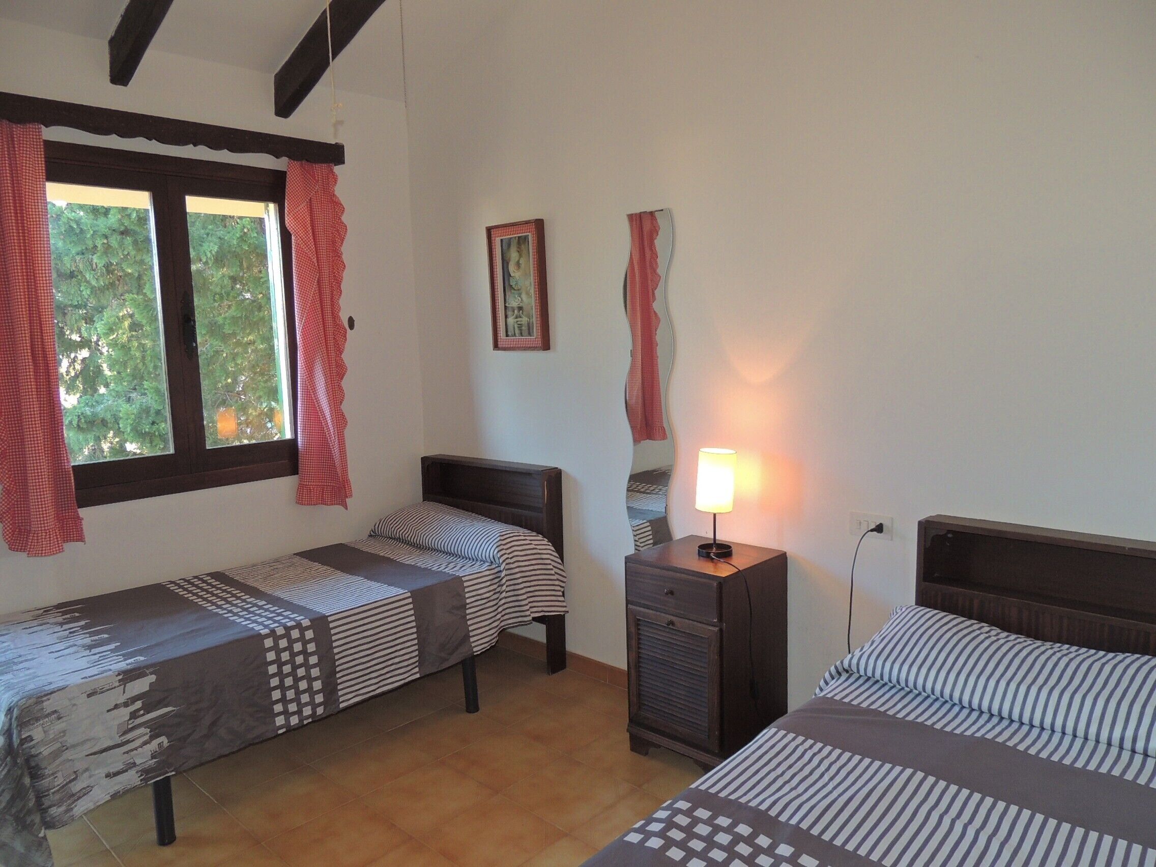 Une chambre avec deux lits simples, une table de chevet avec une lampe, une fenêtre avec des rideaux et un tableau encadré au mur.