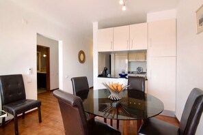 A dining area with a glass table surrounded by chairs, a kitchen in the background, and a door leading to another room.