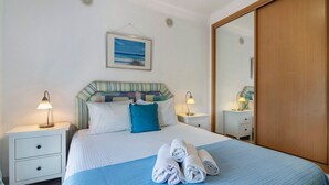 Ein ordentlich bezogenes Bett mit Kissen, einer blauen Tagesdecke und gerollten Handtüchern. Zwei Nachttische mit Lampen, ein Wandbild und ein Holzschrank sind ebenfalls vorhanden.