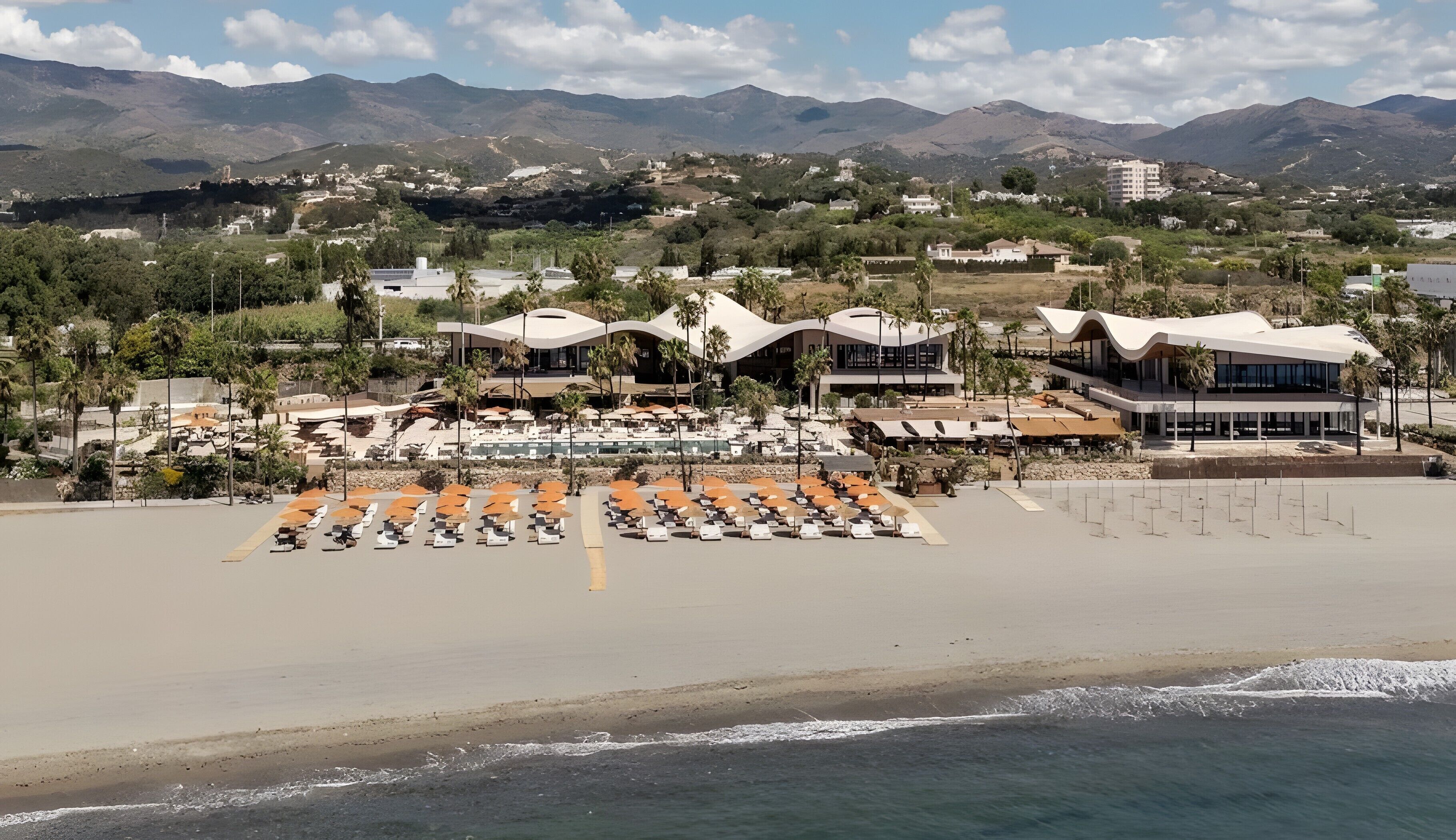 Un hotel frente a la playa con zona de piscina, tumbonas y arquitectura moderna.