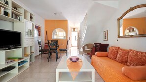A living room with a TV, bookshelf, orange sofa, and a dining area with chairs and a table. - Holiday Home Casa Sarita with Shared Pool & Wi-Fi (Arona)