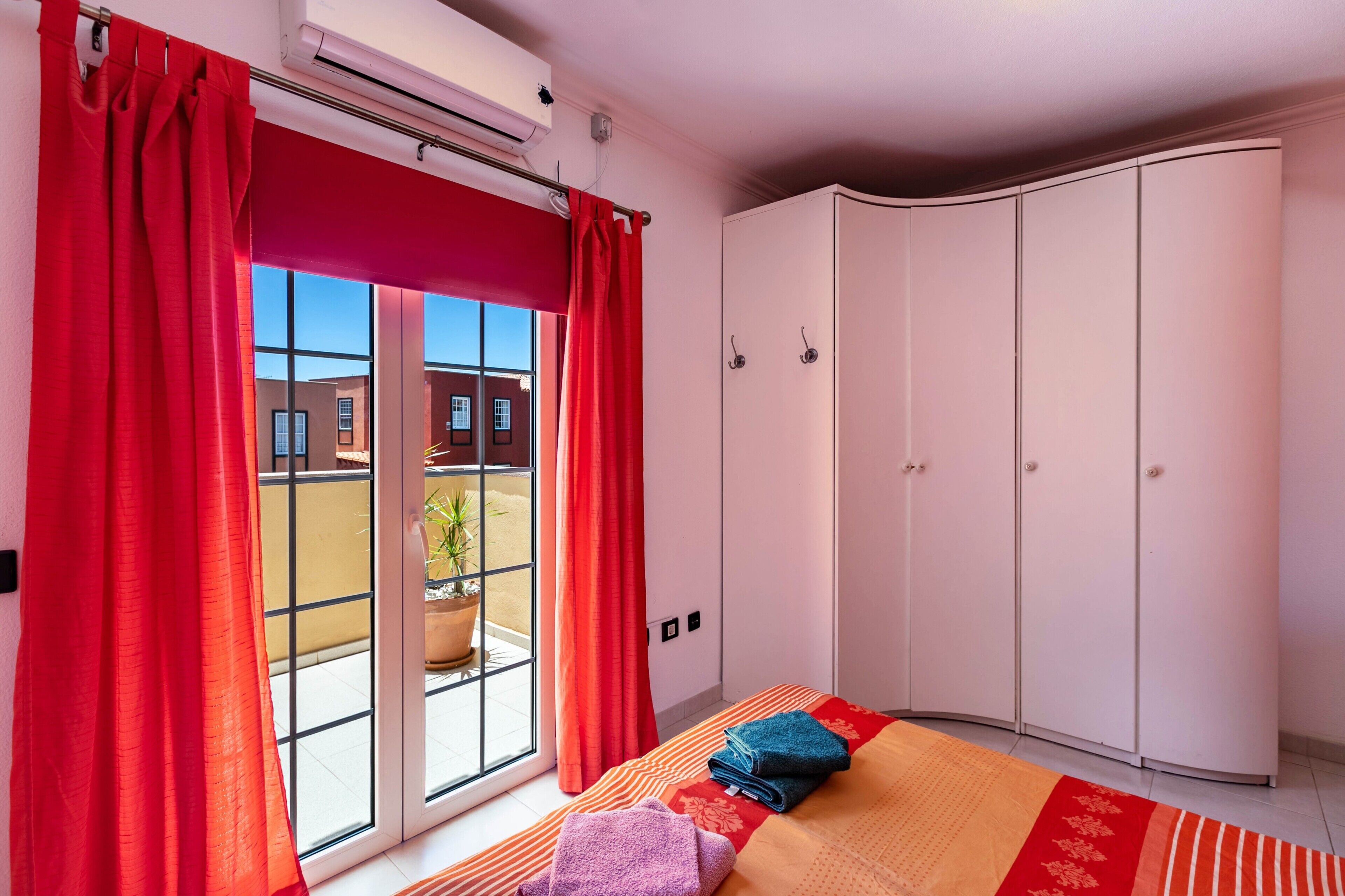 Ein Schlafzimmer mit großem Kleiderschrank, einem Bett mit roter und orangefarbener Bettdecke und einem Fenster mit roten Vorhängen.