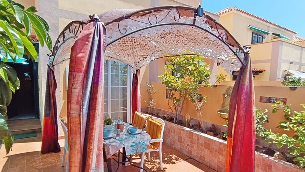Une terrasse extérieure avec une table et des chaises, un espace salon couvert et un jardin agrémenté de plantes et d’un arbre.