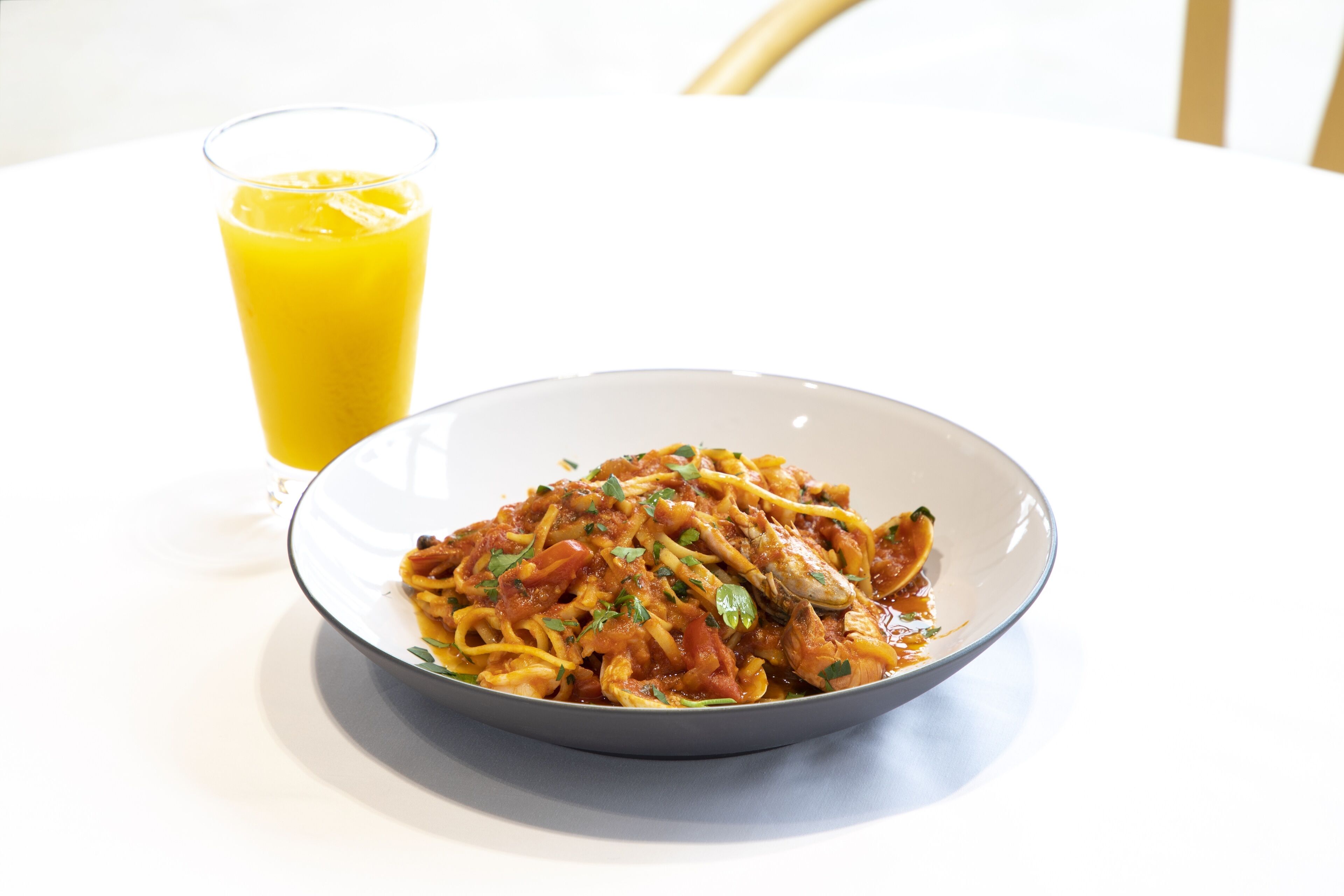 Un plato de espagueti con salsa de tomate y hierbas, y un vaso de jugo de naranja sobre una mesa blanca.