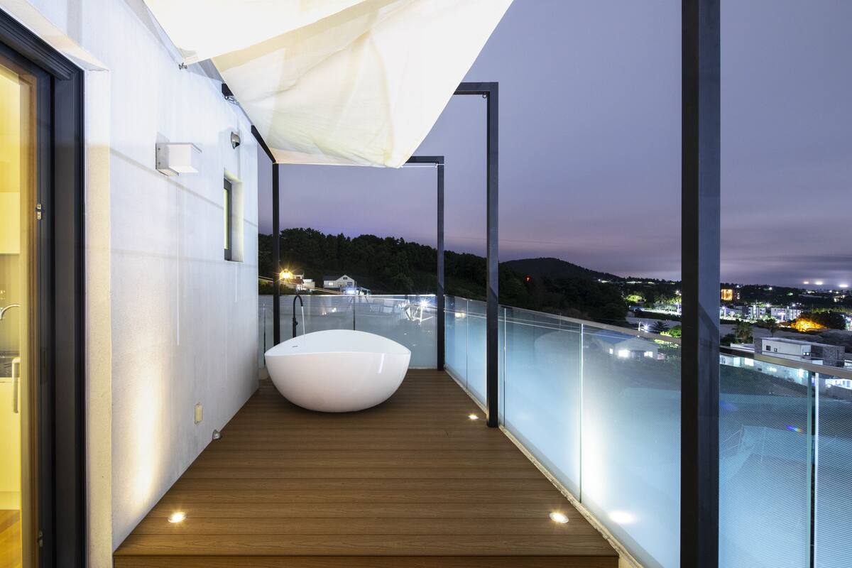 A modern balcony with a wooden floor, a white bathtub, and a glass railing overlooking a cityscape at dusk.