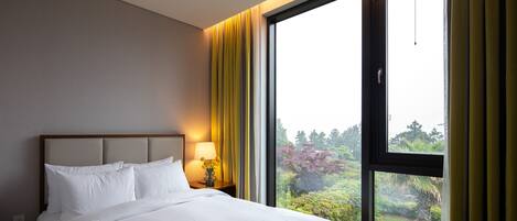 A hotel room with a large window, a bed with white linens, a nightstand with a lamp, and a view of greenery outside.