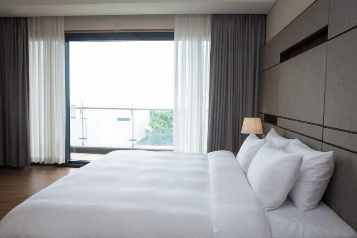 A hotel room with a large bed, a nightstand with a lamp, and a view of the outdoors through a window.