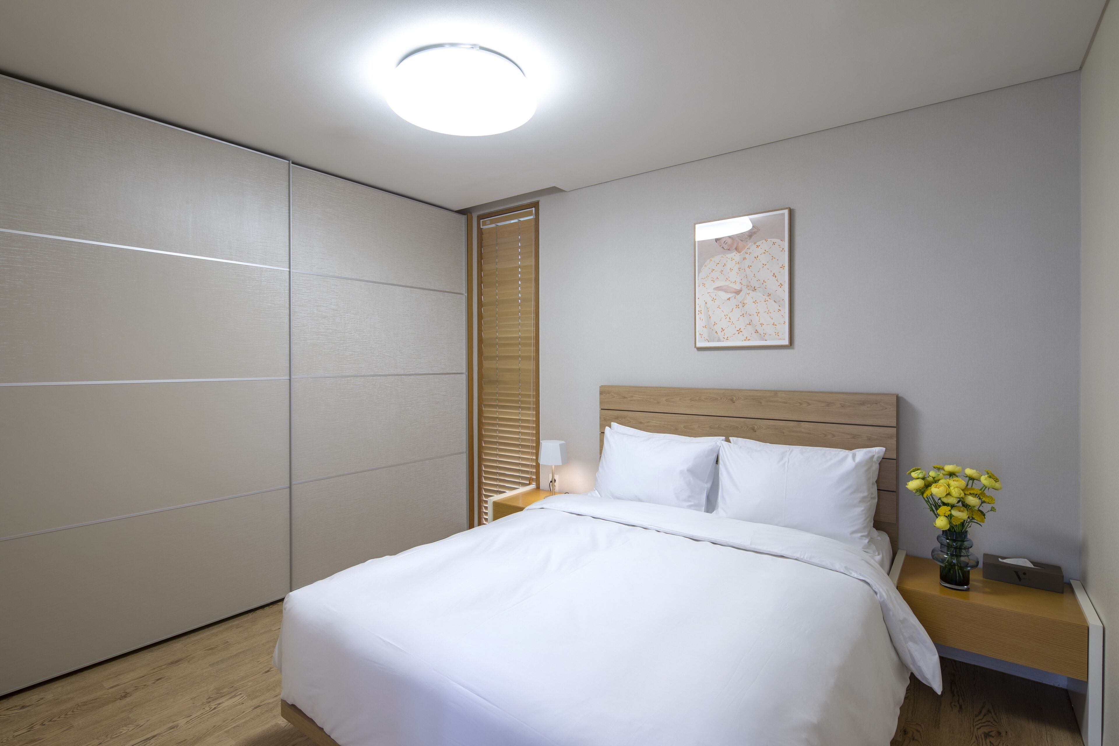 Una cama bien hecha con sábanas blancas, cabecera de madera, mesita de noche con lámpara y un florero con flores amarillas.