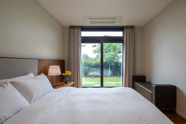 A hotel room with a large bed, a nightstand with a lamp and flowers, a TV, and a sliding glass door to the outside. - Velum Resort Jeju (Seogwipo)