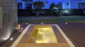 Un área moderna de hidromasaje al aire libre con un muelle de madera y guijarros blancos, rodeada por un seto bajo y vistas despejadas de edificios residenciales al fondo.