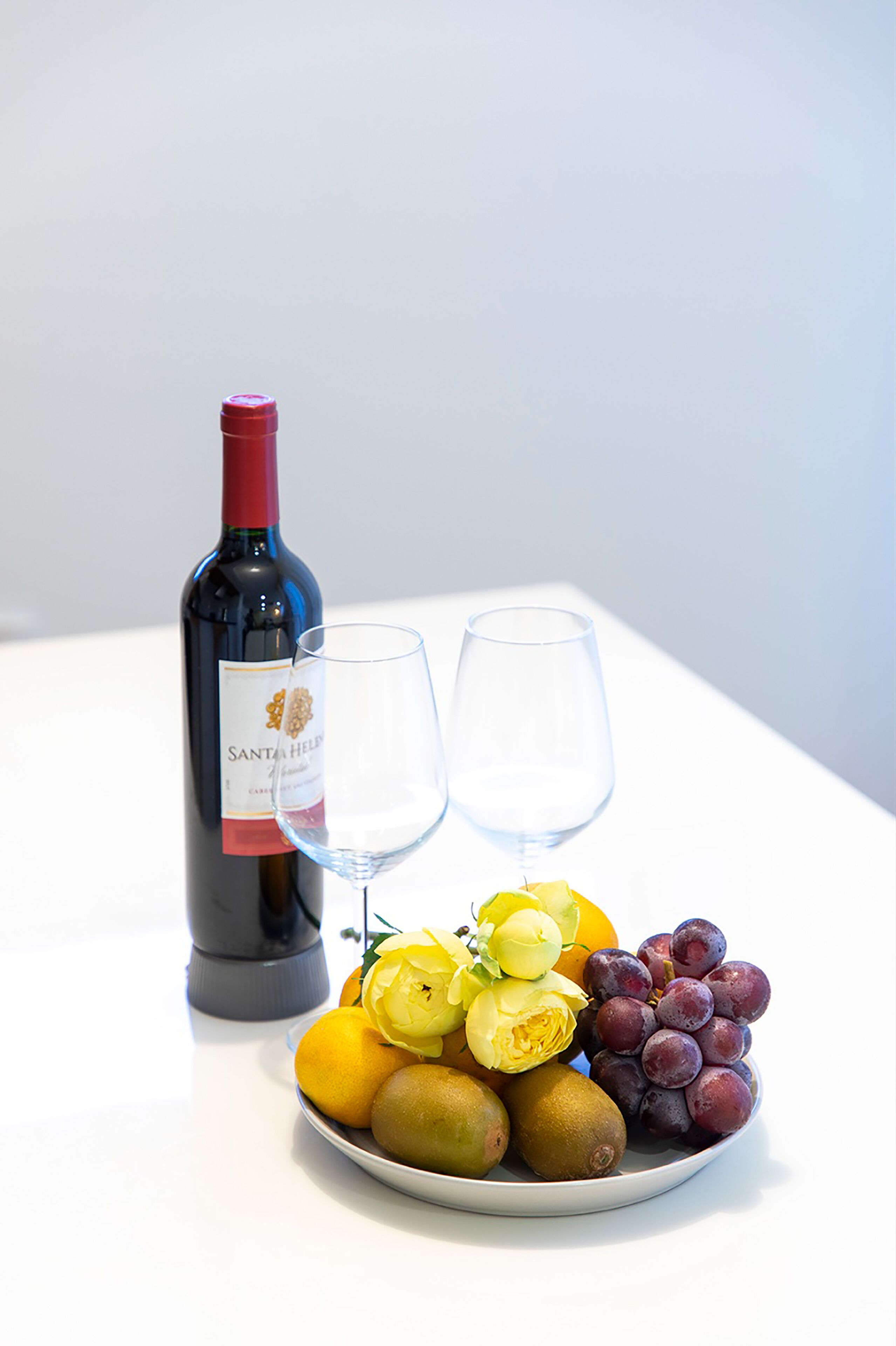 A bottle of wine, two wine glasses, and a plate with fruits and flowers.