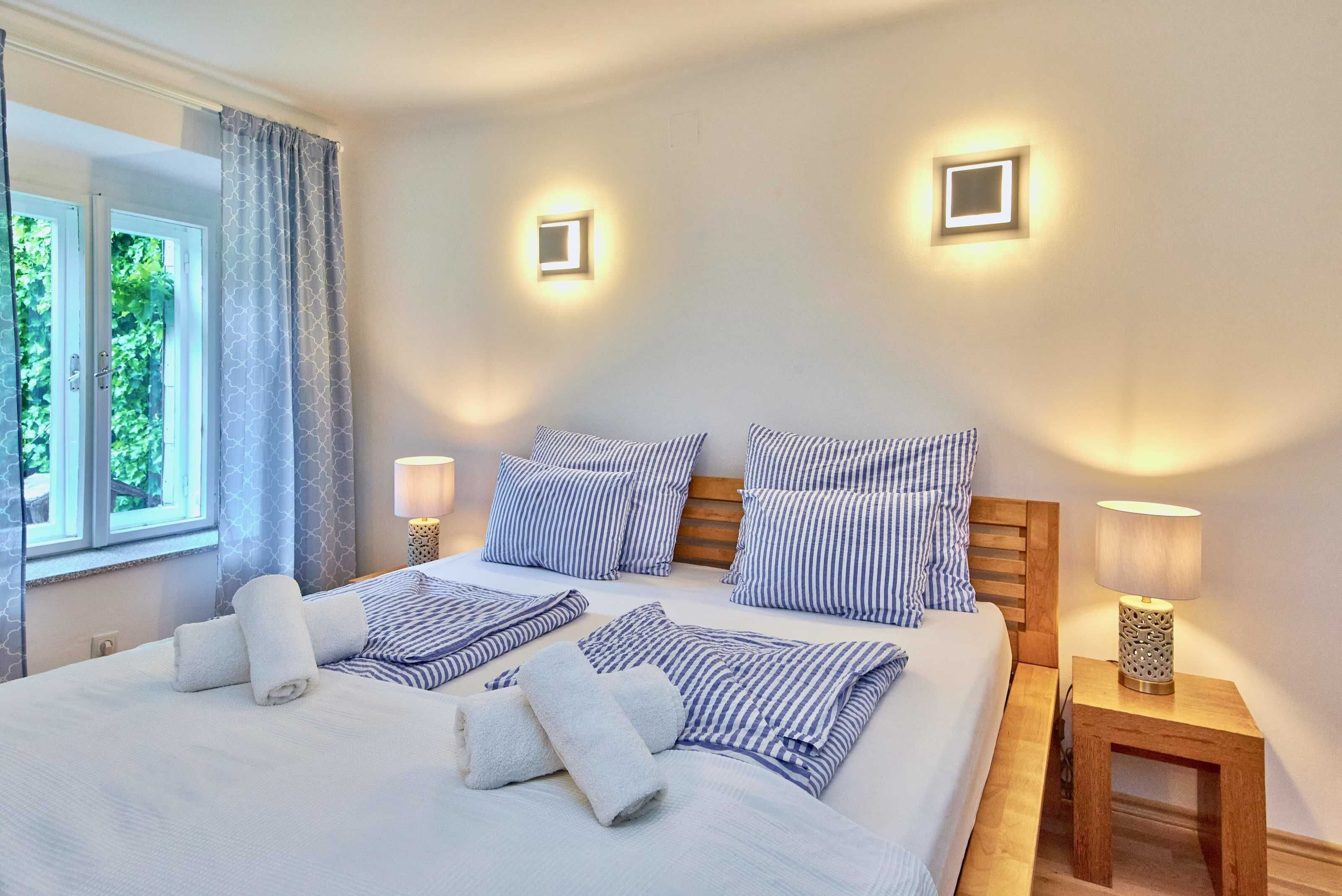 A neatly made bed with striped pillows and towels, two bedside lamps, and a window with curtains.