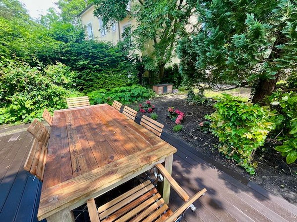 En træterrasse med et bord og stole, omgivet af frodig vegetation og et hus i baggrunden.